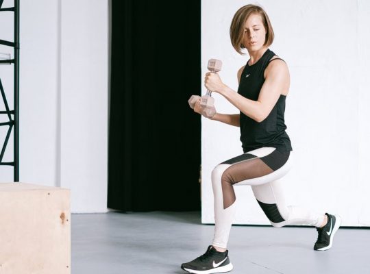 A young woman performing lunges with dumbbells in a modern indoor gym setting.