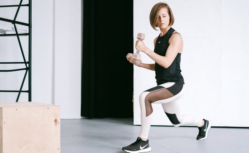 A young woman performing lunges with dumbbells in a modern indoor gym setting.